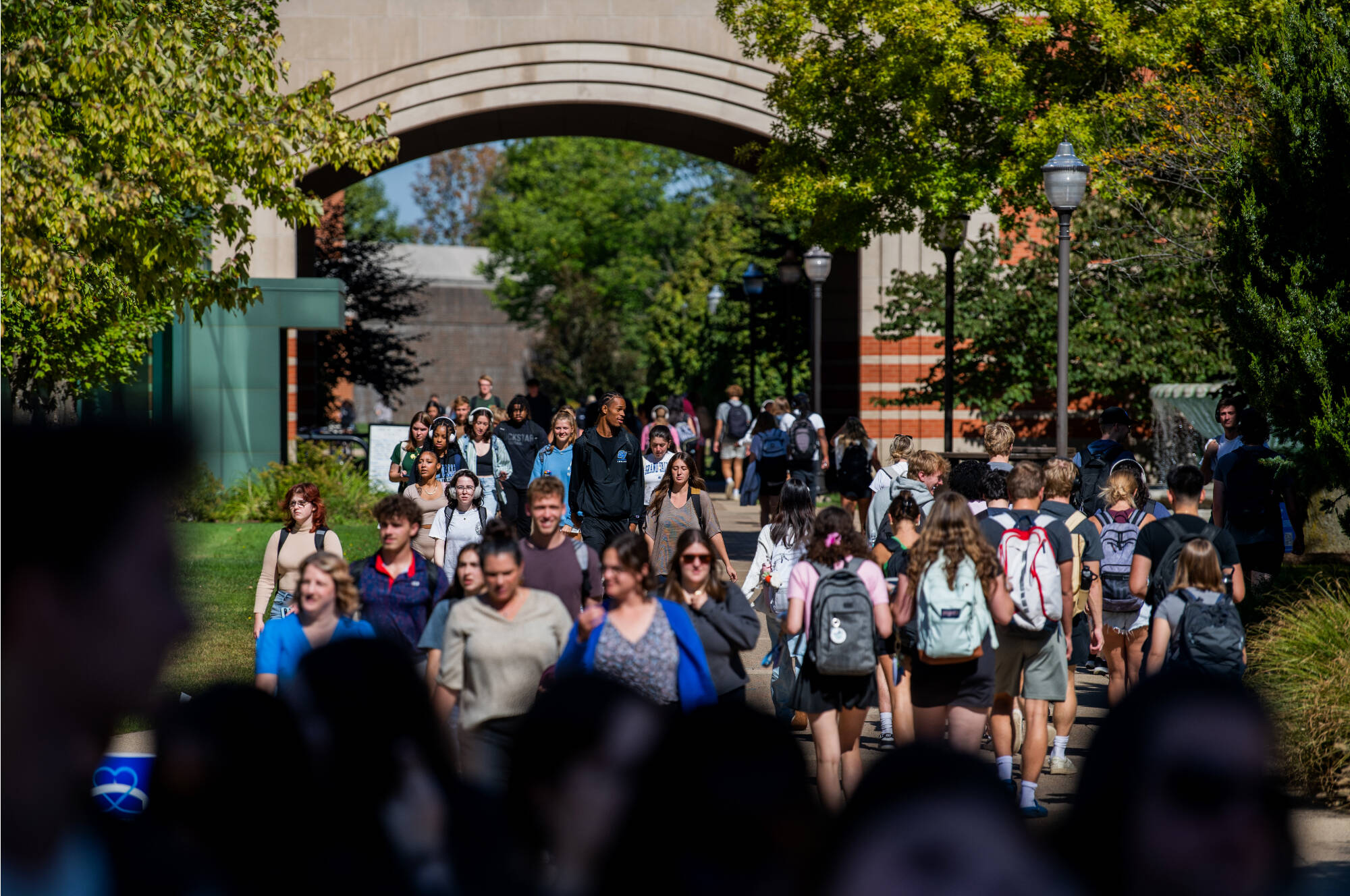 Diverse group of students with backpacks walk along busy campus pathway beneath large archway, with trees, lampposts, and academic buildings visible on sunny day between classes.